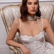 Woman in a white lace dress sitting on an ornate chair with a pearl necklace.