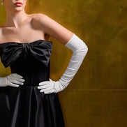 Woman wearing a black dress with a large bow and white gloves against a yellow background