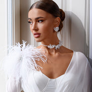 Woman in a white bridal gown with feathered shoulders and jewelry, standing against a neutral wall.
