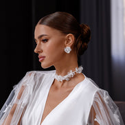 Woman wearing a white wedding dress with floral details and jewelry against a dark curtain background